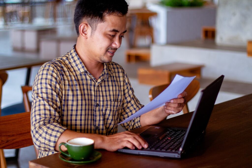 Man sitting at coffee ship table with open laptop, looking at a sheet of paper